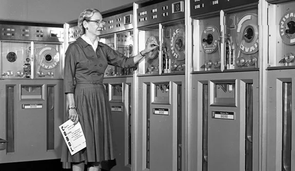 An image of Grace Hopper, one of the pioneers of compiler languages, with old fashioned computers.
